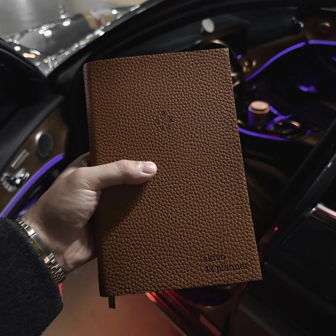 Brown leather AKIVO 4X Planner held beside a car